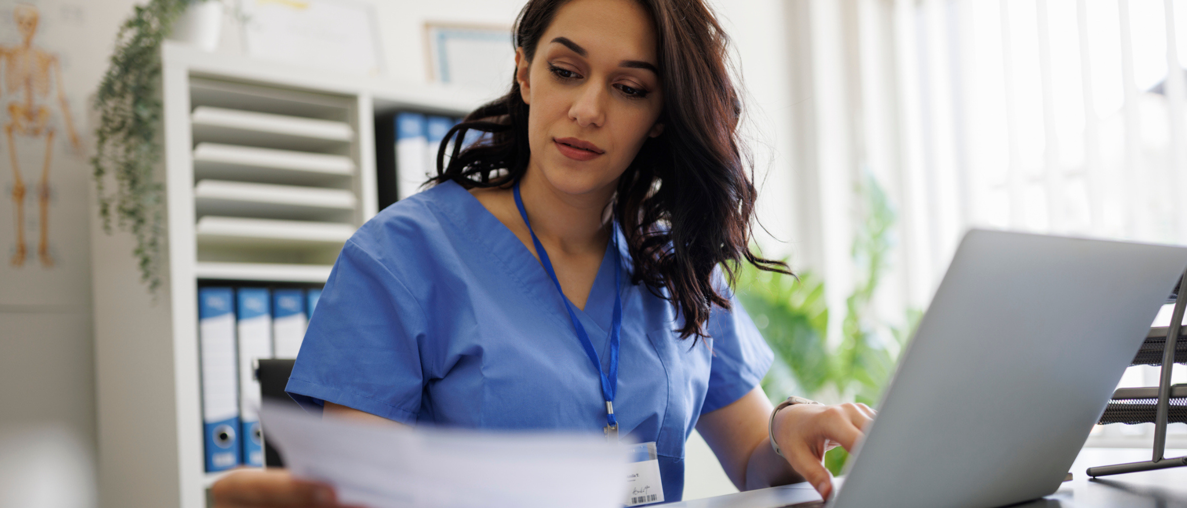 A healthcare worker in blue scrubs reviews a paper document while typing on a laptop in a bright clinic office.