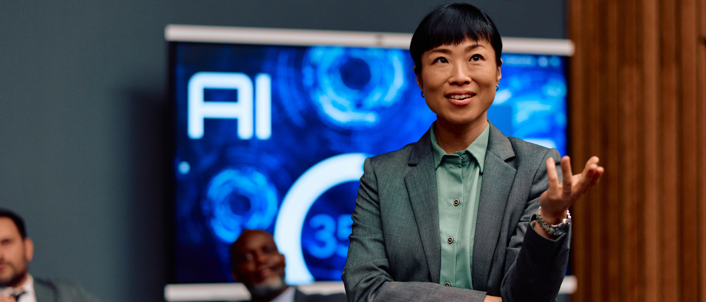 A professional woman in a gray blazer and green shirt speaks to colleagues in a meeting room, gesturing with one hand, in front of a screen displaying “AI” and data graphics.